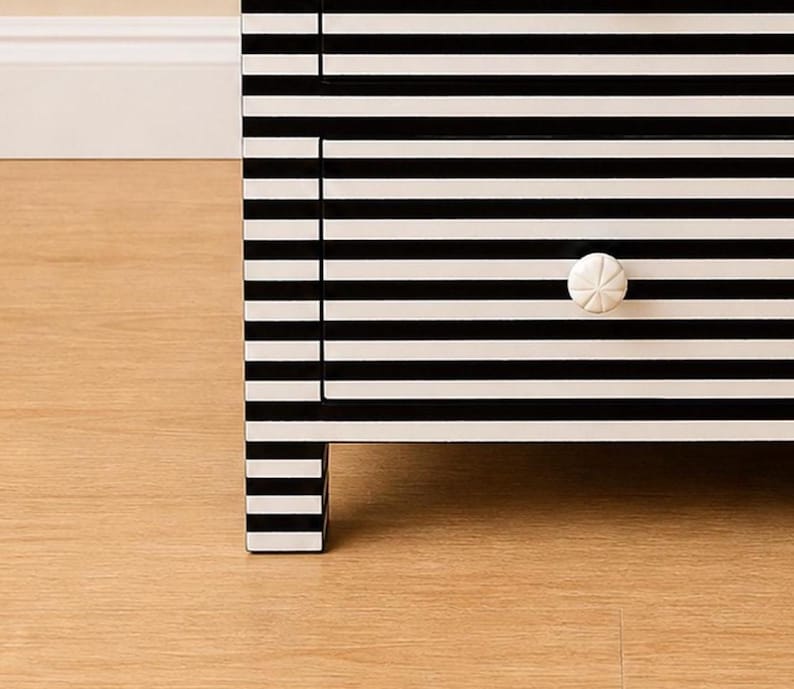 May include: A black and white striped dresser with a single drawer. The drawer has a white, starburst-shaped knob. The dresser is made of wood and has a modern design. The legs of the dresser are also striped.