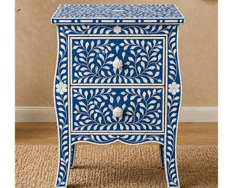 May include: A blue nightstand with two drawers, adorned with white floral and leaf patterns. The nightstand has a curved design and white flower-shaped drawer pulls. The top and sides feature intricate inlaid designs.