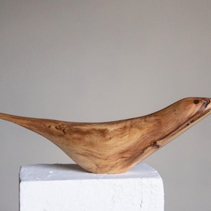 Könnte beinhalten: Eine Holzskulptur eines Vogels mit einem langen, schlanken Schnabel und einem geschwungenen Körper. Die Skulptur ist aus hellbraunem Holz mit dunkelbraunen Streifen.