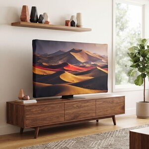 May include: A flat screen television displaying a desert landscape with rolling sand dunes in shades of orange, red, and dark brown, positioned on a long, rectangular walnut wood media console with three cabinet doors and tapered legs.
