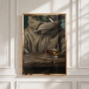 May include: Framed artwork depicting a green alien-like figure in a brown hooded coat, smoking a cigarette, with a glass of whiskey on a wooden table. The artwork is framed in a gold-colored frame.