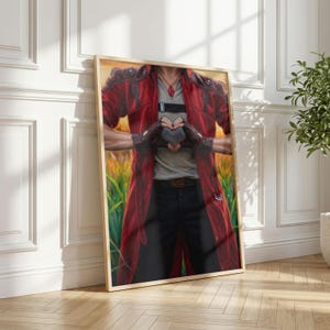 May include: Framed artwork featuring a person in a red coat and black pants, forming a heart shape with their hands. The artwork has a gold frame and is leaning against a white wall. The background shows a field of wheat.