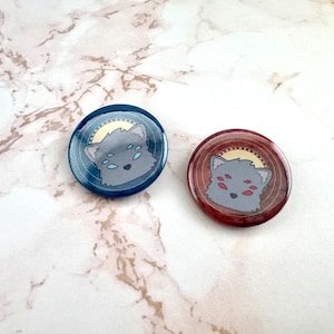 Puede incluir: Dos botones redondos con pasador con un diseño de cabeza de lobo de dibujos animados. El lobo tiene varios ojos y un halo. Un botón tiene un fondo azul y el otro tiene un fondo rojo.