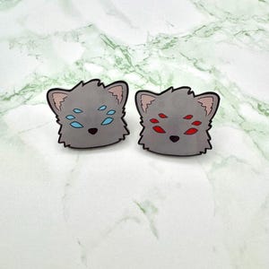 May include: Two gray, cartoon-style cat-like face earrings. One pair has blue eye accents, the other has red eye accents. The earrings have a black outline and are set against a white and green marble background.