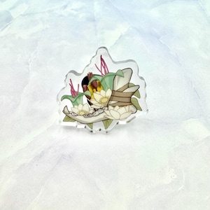 May include: A clear acrylic charm featuring a white skull with green leaves and pink flowers. The skull is surrounded by white flowers and green leaves.