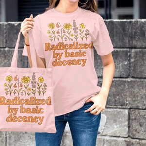 May include: A light pink t-shirt and tote bag with the text "Radicalized by basic decency" in orange. Both items feature a floral design. The person is wearing blue jeans.