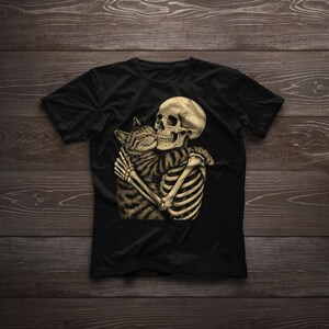 May include: Black t-shirt with a graphic design of a skeleton hugging a tabby cat. The skeleton is depicted with its rib cage and arms visible, holding the cat. The design uses beige and brown tones, contrasting with the black of the shirt.