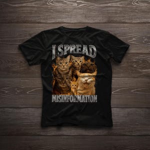 May include: Black t-shirt featuring a graphic of several cats with flames and the text "I SPREAD MISINFORMATION" in silver. The cats have various fur colors, including brown, orange, and black. The shirt is displayed on a wooden surface.