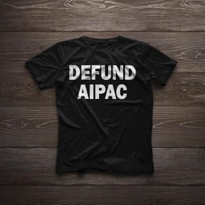 Puede incluir: Camiseta negra con el texto blanco "DEFUND AIPAC" impreso en la parte delantera. La camiseta se muestra sobre una superficie de madera oscura. El texto está en negrita, con una fuente sans-serif.