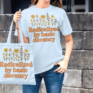 May include: Light blue t-shirt and tote bag with the text "Radicalized by basic decency" in orange. Both items feature a floral design in yellow, orange, and purple. The person is wearing blue jeans.
