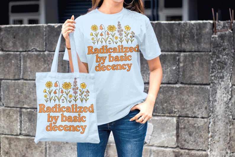 May include: Light blue t-shirt and tote bag with the text "Radicalized by basic decency" in orange. Both items feature a floral design in yellow, orange, and purple. The person is wearing blue jeans.