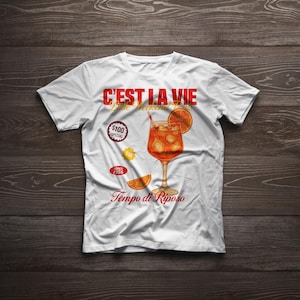 May include: White t-shirt with a colorful graphic of a cocktail, orange slice, and ice cubes. Text includes "C'est La Vie", "Little Weekend Beats", and "Tempo di Riposo". Additional text includes "$100 Special" and "Since 1956".