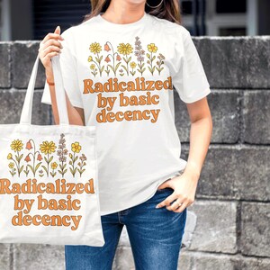 May include: White t-shirt and tote bag with the text "Radicalized by basic decency" in orange lettering. Both items feature a floral design in yellow, orange, and purple. The person is wearing blue jeans.