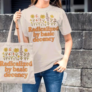 May include: A cream-colored t-shirt and tote bag with the phrase "Radicalized by basic decency" in orange lettering. Both items feature a floral design above the text. The person is wearing blue jeans.