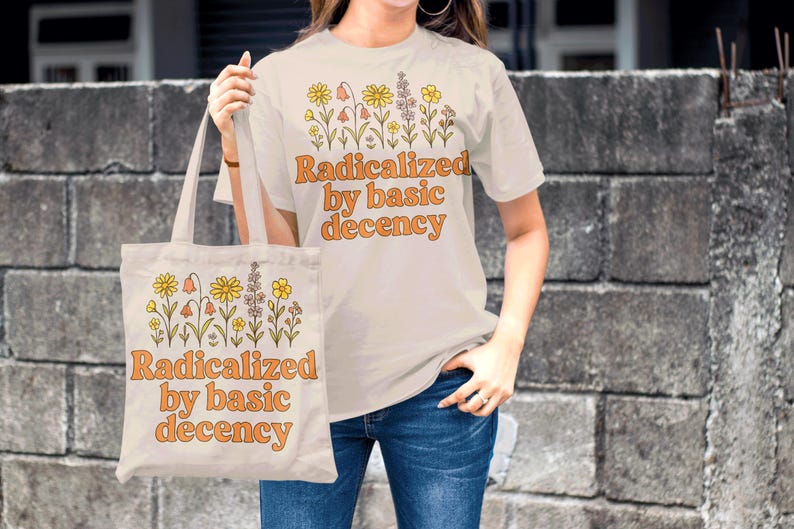 May include: A cream-colored t-shirt and tote bag with the phrase "Radicalized by basic decency" in orange lettering. Both items feature a floral design above the text. The person is wearing blue jeans.