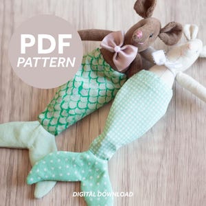 Puede incluir: Dos peluches de sirena hechos a mano. Uno es marrón con un lazo rosa y una cola de escamas verdes. El otro es blanco con un lazo blanco y una cola a cuadros verde claro. El texto "PDF PATTERN" está en un círculo encima de los juguetes. El texto "DESCARGA DIGITAL" está debajo.