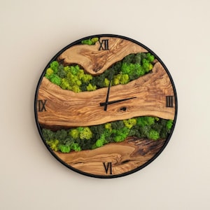 Puede incluir: Reloj de pared redondo con marco de metal negro, con un diseño de vetas de madera natural y detalles de musgo verde. La esfera del reloj tiene números romanos negros y manecillas negras. Una pieza decorativa única.