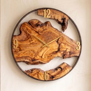 May include: A round wall clock with a natural wood face and a dark metal frame. The clock face features a unique wood grain pattern with gold-colored numerals at 12, 3, 6, and 9 o'clock.