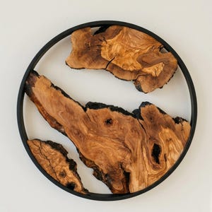 Peut inclure: Décoration murale circulaire en bois avec des motifs de grain de bois naturels bruns et beiges, encadrée dans un cadre en métal noir. Le bois a une forme organique unique, ajoutant une touche rustique et artistique.