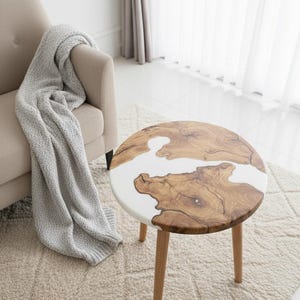 Puede incluir: Una mesa auxiliar redonda con una tapa de vetas de madera y una incrustación de resina blanca. La mesa tiene tres patas de madera y está sobre una alfombra beige. Una manta gris está sobre un sillón beige.
