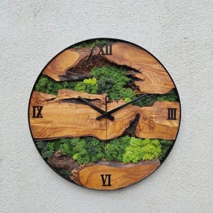 May include: A round wooden clock with Roman numeral markings and a black metal frame. The clock face is decorated with green moss and natural wood.