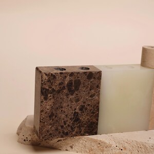 May include: A collection of decorative stone objects. A rectangular brown marble block with two holes sits next to a white rectangular block. A small, cylindrical object is also present. The objects rest on a textured stone base.