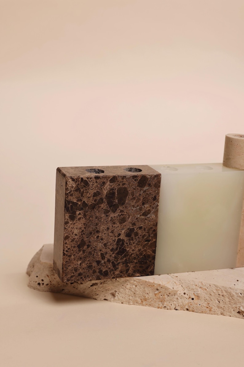 May include: A collection of decorative stone objects. A rectangular brown marble block with two holes sits next to a white rectangular block. A small, cylindrical object is also present. The objects rest on a textured stone base.