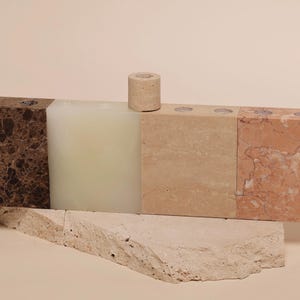 May include: A multi-colored stone candle holder with a cylindrical candle. The holder is composed of various stone blocks in shades of brown, beige, and pink. The candle is a light beige color. The holder sits on a rough-hewn stone base.