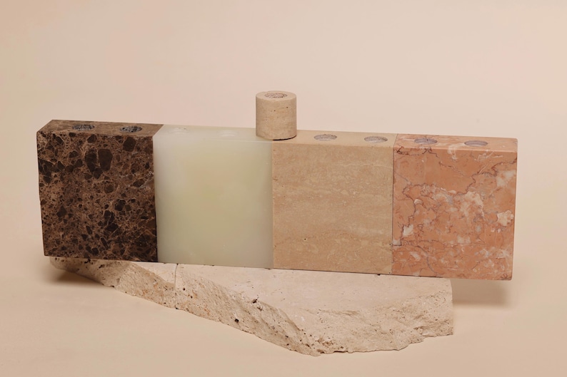 May include: A multi-colored stone candle holder with a cylindrical candle. The holder is composed of various stone blocks in shades of brown, beige, and pink. The candle is a light beige color. The holder sits on a rough-hewn stone base.
