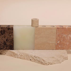 May include: A multi-colored stone candle holder with four sections, including brown, off-white, beige, and reddish-brown. A small, cylindrical candle holder sits on top. The candle holder rests on a light-colored, textured stone base.