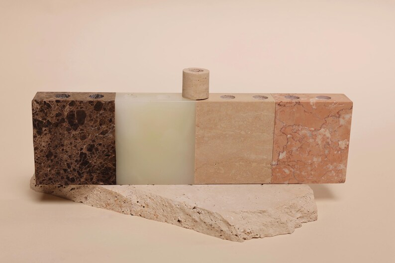 May include: A multi-colored stone candle holder with four sections, including brown, off-white, beige, and reddish-brown. A small, cylindrical candle holder sits on top. The candle holder rests on a light-colored, textured stone base.