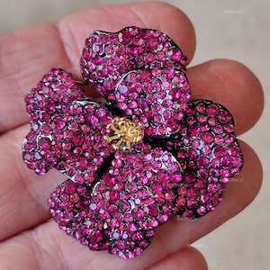 May include: A flower-shaped brooch with a dark metal outline. The brooch is covered in small, sparkling magenta crystals. The center of the flower has a gold-coloured, textured design. A beautiful piece of jewellery.
