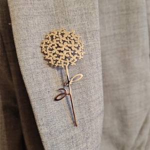 Pode incluir: Um broche de metal dourado em forma de flor com um design delicado e intrincado. A flor tem uma forma redonda e é composta por muitas pétalas pequenas que se sobrepõem. O caule é fino e tem uma única folha.