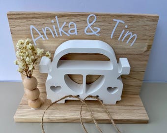 Peut inclure: Décoration de mariage en bois avec une découpe en forme de voiture blanche, des détails en forme de cœur et les noms "Anika & Tim" en blanc. Sur un fond en bois clair, avec un petit vase de fleurs séchées et de la ficelle.