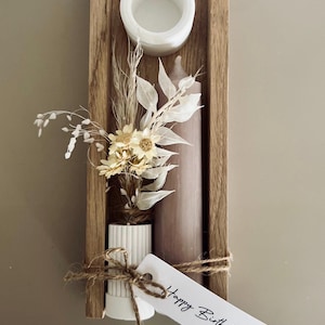 Könnte beinhalten: Holzkerzenhalter mit einer weißen Keramikschale, einer hohen braunen Kerze und einer kleinen Vase mit Trockenblumen. Ein Etikett mit "Happy Birthday" ist mit Bindfaden befestigt. Die Anordnung hat eine neutrale Farbpalette.