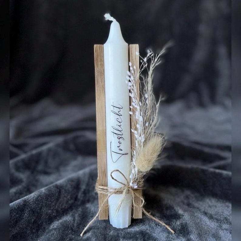 May include: A white taper candle featuring the black script text Trostlicht, held between two vertical light wood slats. The candle is decorated with a small bundle of dried white and tan grasses tied with a thin brown twine bow at the base.