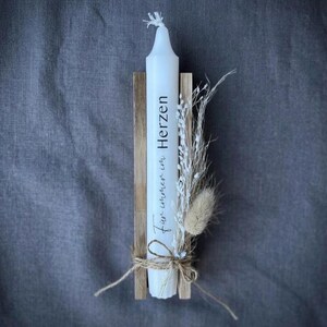 May include: A white taper candle featuring the black text "F&uuml;r immer im Herzen" printed vertically. The candle is secured between two light brown wooden slats and decorated with a bundle of dried white flowers and a small beige grass plume.