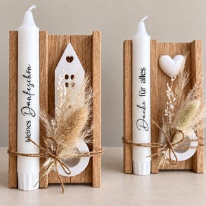Op de afbeelding: Twee witte kaarsen in houten houders met gedroogd gras en touw. De linkerkaars bevat de tekst "kleines Dankeschön" en een wit huisje. De rechterkaars bevat de tekst "Danke für alles" en een wit hartje als decoratie.