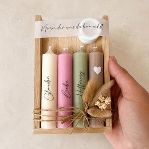 Könnte beinhalten: Ein Holztablett mit vier Stumpenkerzen in Creme, Rosa, Salbeigrün und Taupe. Die Kerzen tragen die deutschen Wörter Glaube, Liebe und Hoffnung, mit einem weißen Herz auf der taupefarbenen Kerze. Ein Band trägt die Aufschrift Nimm dir was du brauchst.