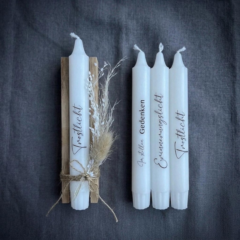 May include: Four white taper candles with black text. One candle is attached to a rectangular wooden block with twine and dried grasses. The candles feature the German words Trostlicht, Im stillen Gedenken, and Erinnerungslicht printed in black script.