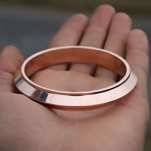 May include: A wide, copper-colored bangle bracelet with a smooth, polished finish. The bracelet is thick and has a rounded, slightly flared edge.