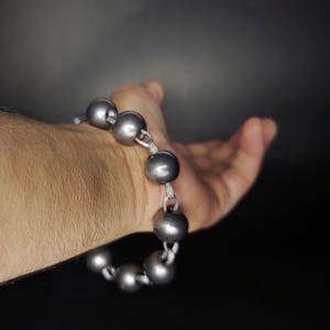 K&ouml;nnte beinhalten: Ein silberfarbenes Armband mit gro&szlig;en, runden Perlen. Die Perlen sind mit einer silbernen Kette verbunden. Das Armband wird an einem Handgelenk getragen, mit offener Hand im Hintergrund. Der Hintergrund ist dunkelgrau.