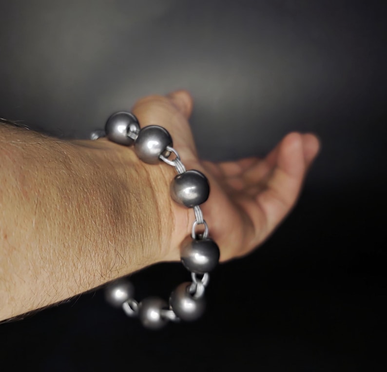 K&ouml;nnte beinhalten: Ein silberfarbenes Armband mit gro&szlig;en, runden Perlen. Die Perlen sind mit einer silbernen Kette verbunden. Das Armband wird an einem Handgelenk getragen, mit offener Hand im Hintergrund. Der Hintergrund ist dunkelgrau.