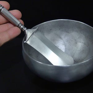 May include: A silver-toned ice cream scoop rests inside a matching bowl. The scoop has a rectangular blade and a faceted handle. The bowl is round with a matte finish. The background is black.