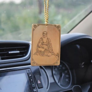 Puede incluir: Encanto de coche dorado con una figura religiosa con barba y turbante. El encanto está colgado de una cadena dorada y tiene la forma de un rectángulo con un borde decorativo.