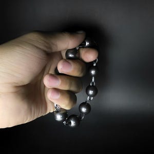 K&ouml;nnte beinhalten: Ein dunkelgraues Perlenarmband mit gro&szlig;en, runden Perlen. Die Perlen sind mit silberfarbenen Metallgliedern aufgef&auml;delt. Das Armband wird in einer Hand vor dunklem Hintergrund gehalten.