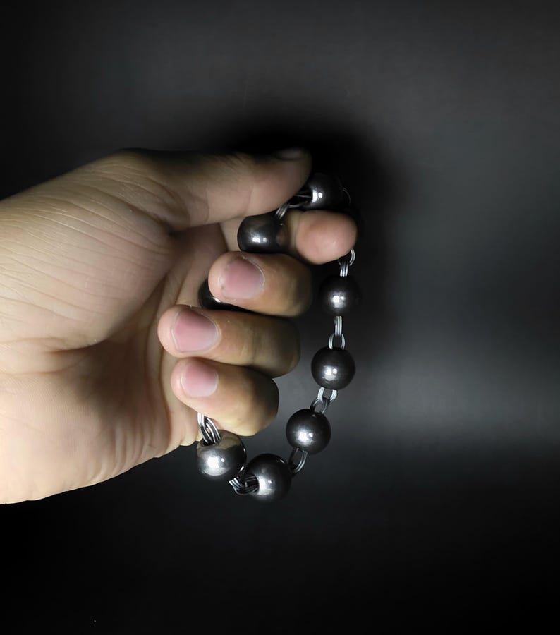 K&ouml;nnte beinhalten: Ein dunkelgraues Perlenarmband mit gro&szlig;en, runden Perlen. Die Perlen sind mit silberfarbenen Metallgliedern aufgef&auml;delt. Das Armband wird in einer Hand vor dunklem Hintergrund gehalten.