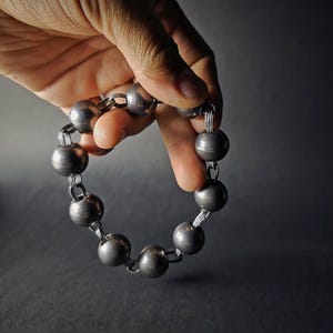 K&ouml;nnte beinhalten: Ein silberfarbenes Armband mit gro&szlig;en, runden Kugeln, die durch kleinere Glieder verbunden sind. Das Armband wird vor einem dunklen Hintergrund gehalten, um seinen metallischen Glanz und sein schlichtes Design hervorzuheben. Die Kugeln haben einen Durchmesser von etwa 2,5 cm.