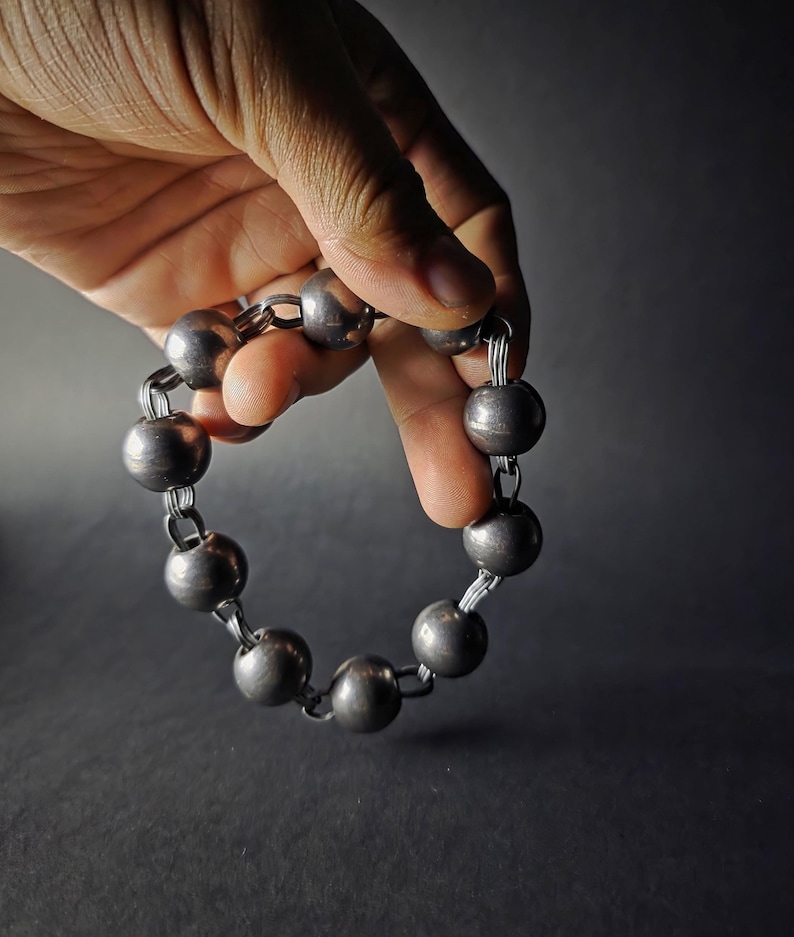K&ouml;nnte beinhalten: Ein silberfarbenes Armband mit gro&szlig;en, runden Kugeln, die durch kleinere Glieder verbunden sind. Das Armband wird vor einem dunklen Hintergrund gehalten, um seinen metallischen Glanz und sein schlichtes Design hervorzuheben. Die Kugeln haben einen Durchmesser von etwa 2,5 cm.