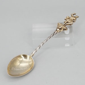 Puede incluir: Una cucharita de plata con mango retorcido y una figura de una mujer con falda escocesa en la parte superior. La cuchara está grabada con la palabra "Edimburgo".